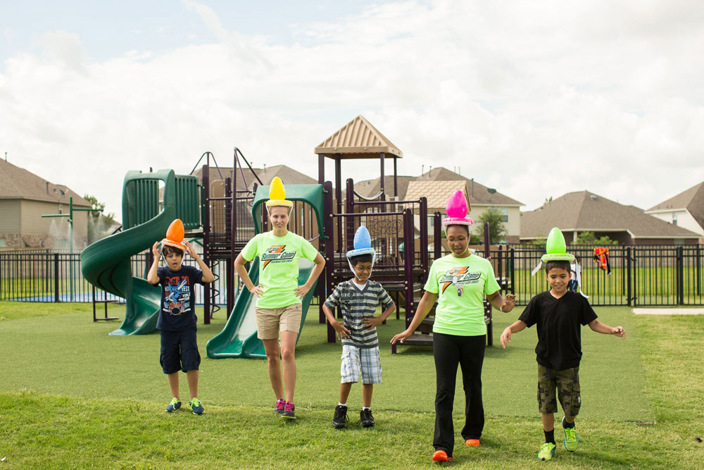 Children and adults wearing colorful cone hats walk on a grassy playground with slides and climbing equipment in the background, enjoying the vibrant atmosphere of an early childhood education franchise.