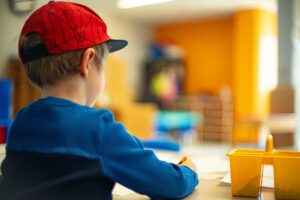 A child draws at a table while wearing a red hat and blue shirt.